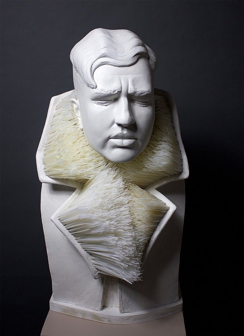 Plaster sculpture of a person with a furrowed brow, a sorrowful expression, and short, styled curly hair, wearing a garment with a collar made of zip ties which imitates an anemone.