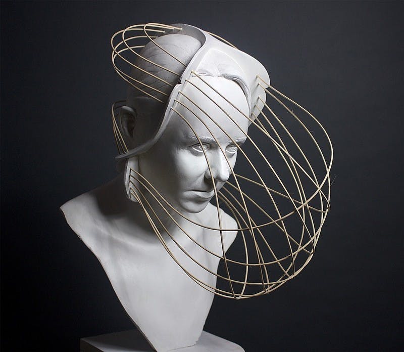 Plaster sculpture of a person with short hair covered by a three-sided headband staring offscreen stoically, with a wicker net protruding from their headband and wrapping around their face and head.